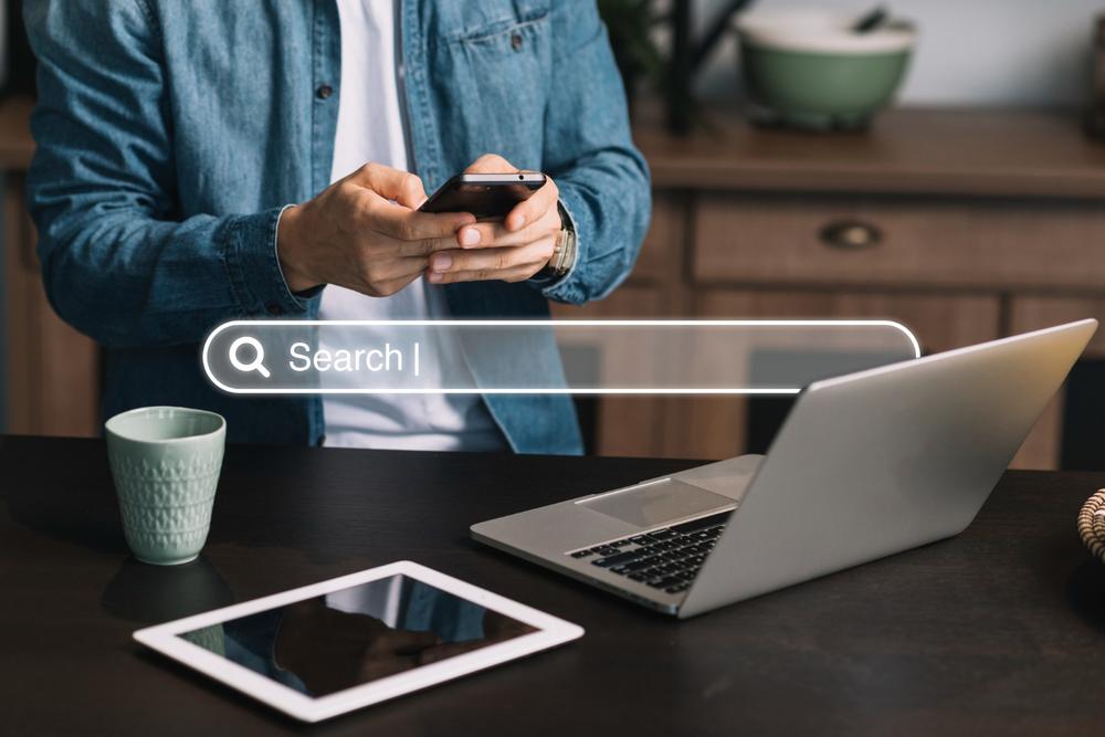 barre de recherche avec en fond un homme tenant un teléphone devant son ordinateur et une tablette